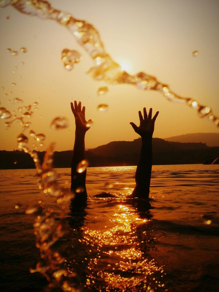 Silhouetted hands reach skyward in a golden sunset, reflecting on rippling water.